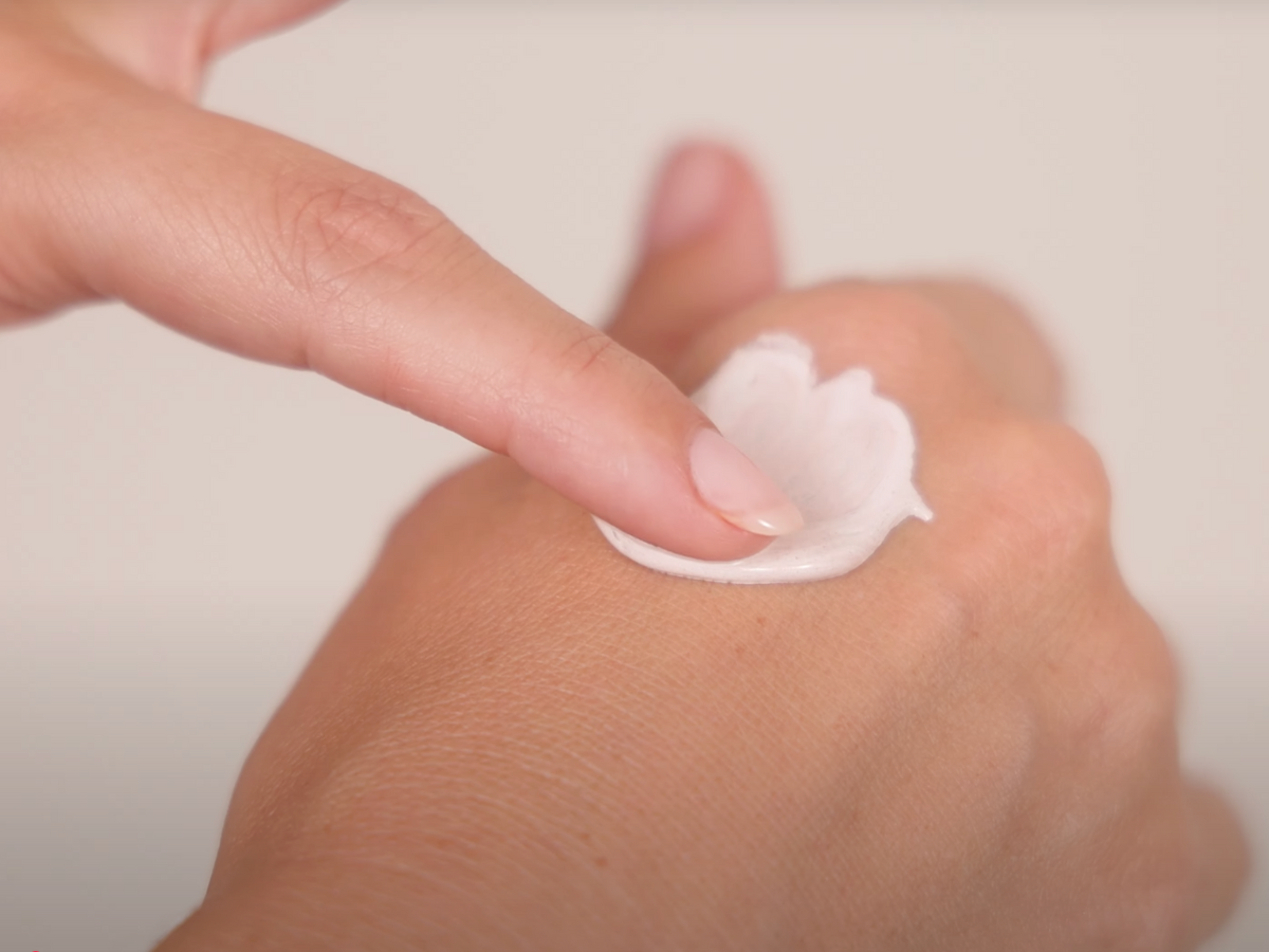 Hand applying a small amount of cream to another hand against a neutral background