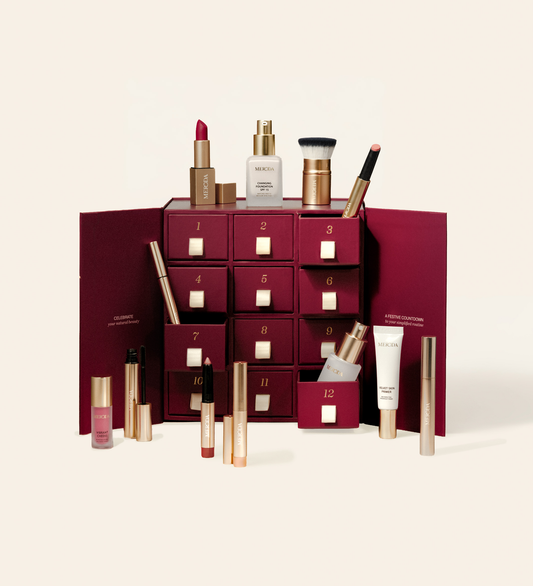 Calendario de Adviento 12 Days of Effortless Beauty con caja burdeos de cajones y productos de maquillaje surtidos sobre fondo crema.